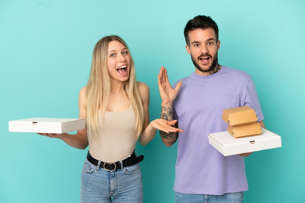 Coppia in possesso di pizze e hamburger su sfondo blu isolato con espressione facciale sorpresa e scioccata