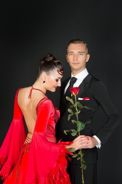 Coppia innamorata del fiore di rosa su sfondo scuro. Donna in abito rosso e uomo in smoking nero, moda. Festa di San Valentino. Amore, proposta, appuntamento. Moda, bellezza, stile.