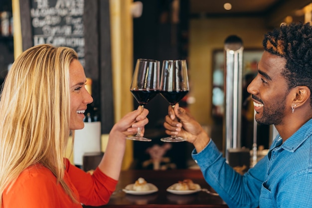 Coppia interrazziale sorridente mentre brindano con bicchieri di vino rosso in un ristorante