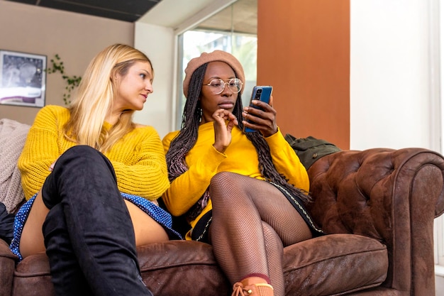 Coppia lesbica di diversa razza e colore della pelle seduta sul divano guardando il cellulare