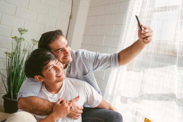 Coppia LGBT caucasica e asiatica che si abbraccia da dietro seduta e utilizza uno smartphone insieme a casa Diversità delle relazioni LGBT Un concetto di convivenza di una coppia gay
