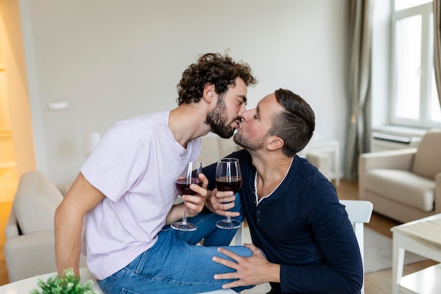 Coppia LGBTQ che si abbracciano e bevono vino al chiuso Due romantici giovani amanti maschi che si guardano l'un l'altro seduti insieme nel loro soggiorno Giovane coppia gay che è romantica a casa