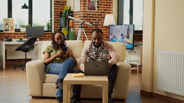 Coppia moderna afroamericana che si rilassa a casa sul divano, scorrendo l'app dei social media sullo smartphone. Godersi attività ricreative e ridere insieme, ricreazione in soggiorno.