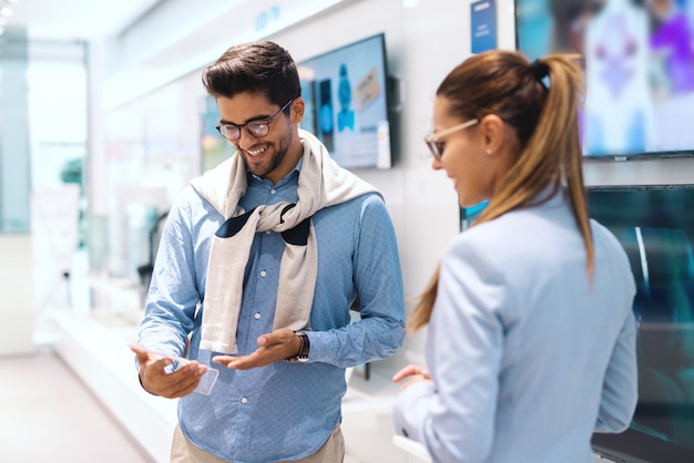 Coppia multiculturale carina sorridente in piedi nel negozio di tecnologia e al prezzo per la nuova televisione che vogliono acquistare