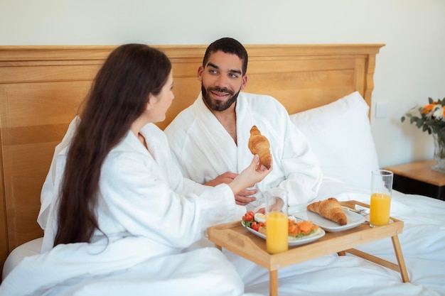 Coppia multiculturale che fa colazione romantica a letto mangiando croissant al coperto