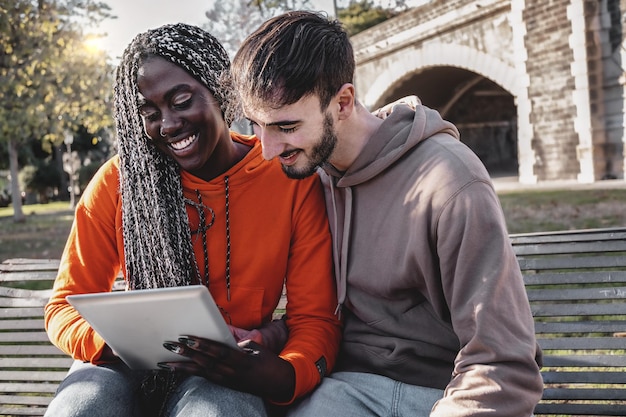 Coppia multietnica di giovani che utilizzano un dispositivo tablet digitale che naviga nei contenuti seduti all'aperto nel parco su una panchina concetto di stile di vita di coppia birazziale che si diverte usando la tecnologia