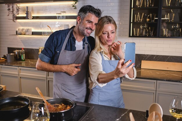 Coppia multirazziale positiva in grembiuli in piedi vicino alla stufa elettrica e prendendo autoritratto sul telefono cellulare mentre si cucina il cibo insieme a casa