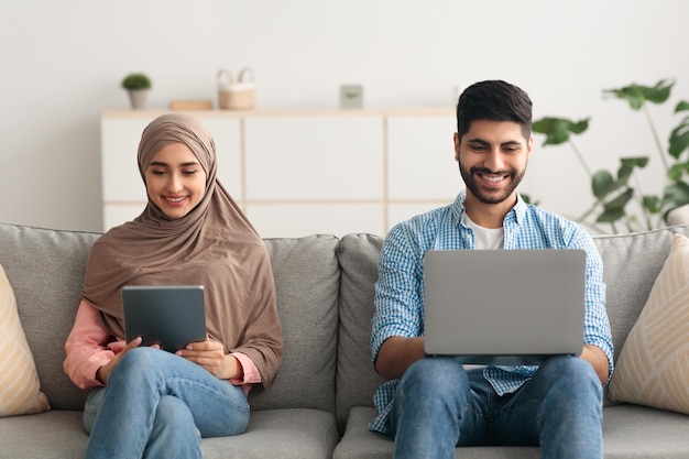 Coppia musulmana che naviga in internet su laptop e tablet digitale all'interno