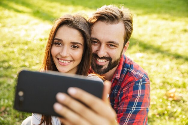 coppia ottimista uomo e donna vestiti in abbigliamento casual che scattano foto selfie sul cellulare mentre riposano nel parco verde green