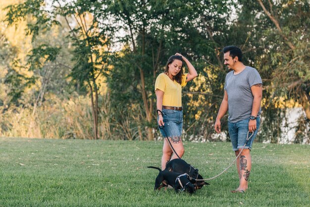Coppia parlando tra loro a piedi con i loro due cani in un parco. Coppia felice parlando con vestiti estivi in una giornata di sole nel parco con i loro due cuccioli.