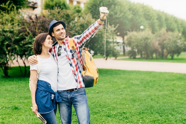 Coppia romantica in amore indossando abiti casual, mentre in piedi sull'erba verde, abbracciati e facendo selfie, sorridendo piacevolmente nella fotocamera. Giovane maschio e femmina a visitare la città