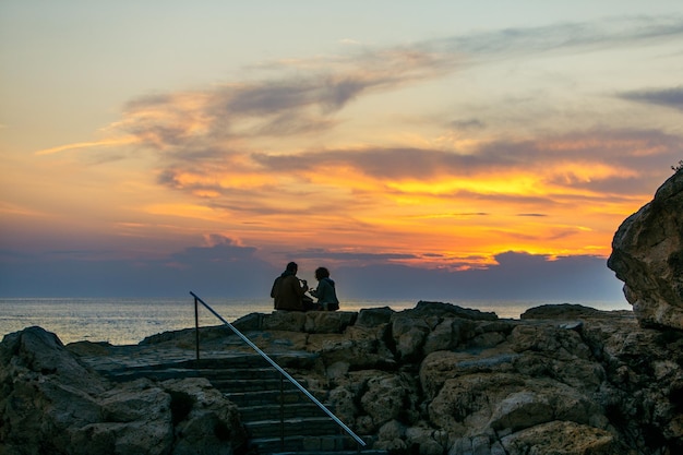 Coppia seduta sulla scogliera rocciosa guardando tramonto romantico appuntamento vacanze estive