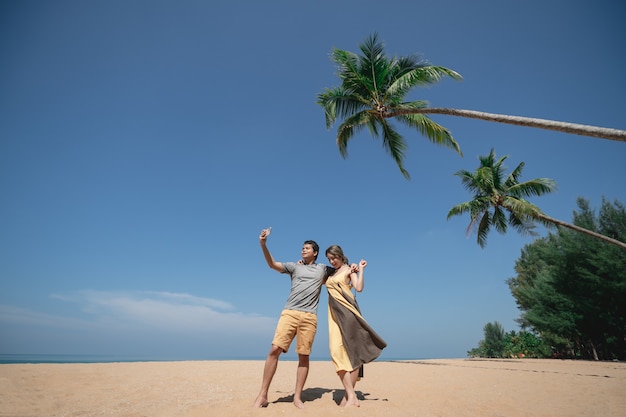 Coppia selfie sotto la palma da cocco sulla spiaggia sabbiosa con cielo blu.