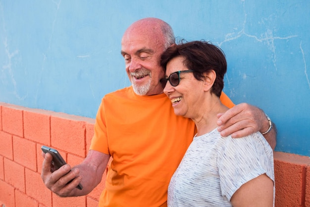 Coppia senior abbracciata e sorridente in piedi contro un muro e guardando il telefono cellulare Felicità e momenti positivi