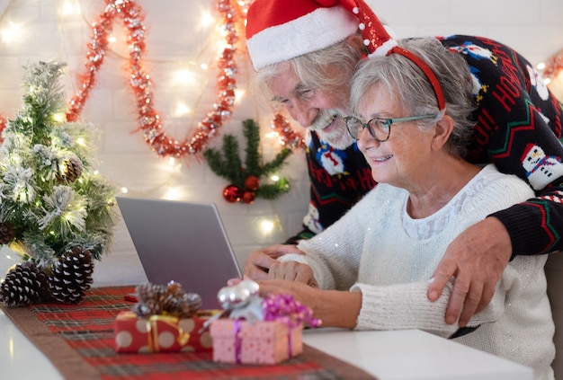 Coppia senior allegra che si abbraccia a Natale più dolce e cappello da Babbo Natale mentre si utilizza il computer portatile