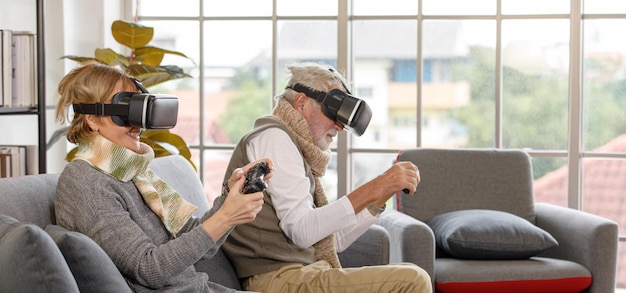 Coppia senior in cuffie VR seduti sul divano e utilizzando i gamepad per giocare a videogiochi contro la finestra nel soggiorno di casa.