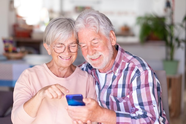 Coppia senior in videochiamata tramite telefono cellulare Bella coppia di anziani gioiosa e sorridente seduta a casa essendo di ottimo umore tenendo lo smartphone