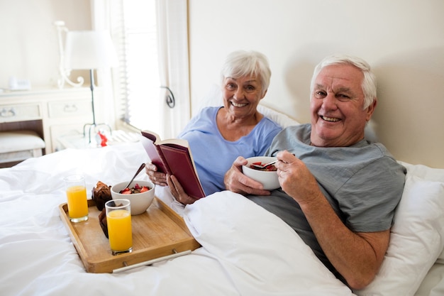 Coppia senior leggendo un libro mentre si fa colazione in camera da letto a casa