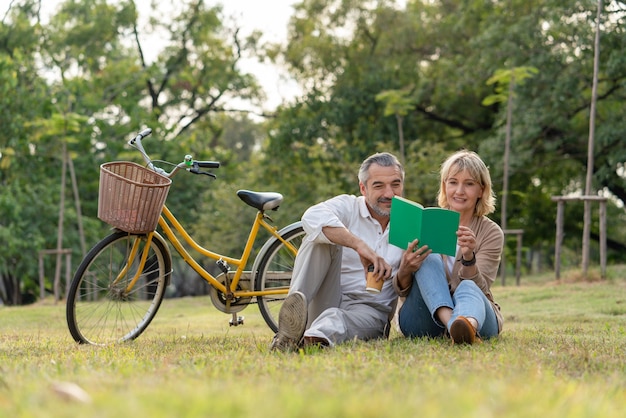 Coppia senior sedersi sull'erba rilassandosi e leggendo un libro insieme dopo aver guidato la bicicletta nel parco