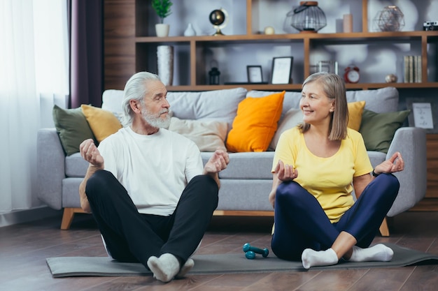 Coppia senior uomo e donna insieme a casa praticando yoga meditando seduti sul pavimento nella posizione del loto
