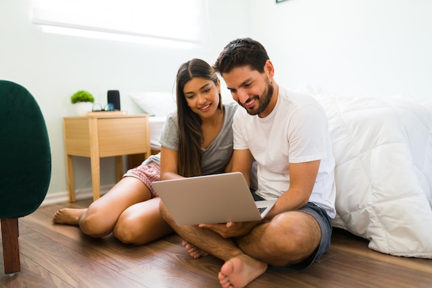 Coppia sorridente che fa un po' di shopping online e cerca prodotti sul proprio laptop mentre è seduta nella loro camera da letto