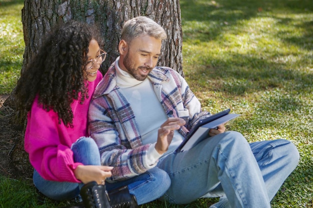 Coppia sorridente e felice seduta sull'erba nel parco guardando lo schermo del tablet Utilizzando la tecnologia moderna