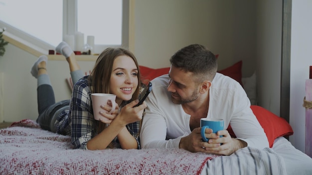 Coppia sorridente guardando la TV sdraiata a letto e bevendo caffè a casa al mattino