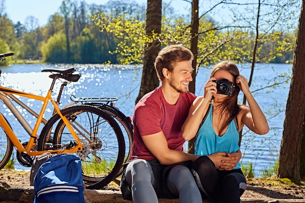 Coppia sportiva positiva che si rilassa vicino al fiume dopo un giro in bicicletta. Femmina che scatta foto su una fotocamera digitale compatta.