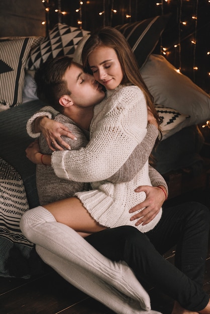 Coppia sul letto in camera da letto. Interno scuro. Baci e abbracci. Essere innamorato. Maglione bianco e calze alte. Incontro romantico. Un appuntamento per gli amanti. Tenerezza.