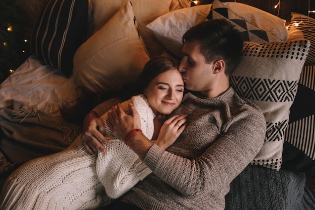 Coppia sul letto in camera da letto. Interno scuro. Capodanno e Natale. Baci e abbracci. Amore. Maglione bianco e calze alte. Incontro romantico. Un appuntamento per gli amanti. Filo rosso al polso.