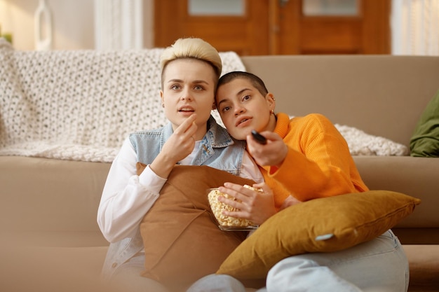 Coppie amorose allegre della donna lesbica che mangiano popcorn che guardano film o show televisivo nel salotto di casa