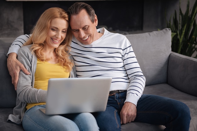 Coppie amorose positive allegre che si siedono sul divano a casa e utilizzano gadget elettronici durante la navigazione in Internet e la condivisione di gioia