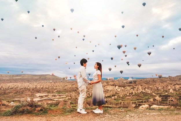 Coppie asiatiche che guardano le mongolfiere variopinte che sorvolano la valle a Cappadocia, Turchia