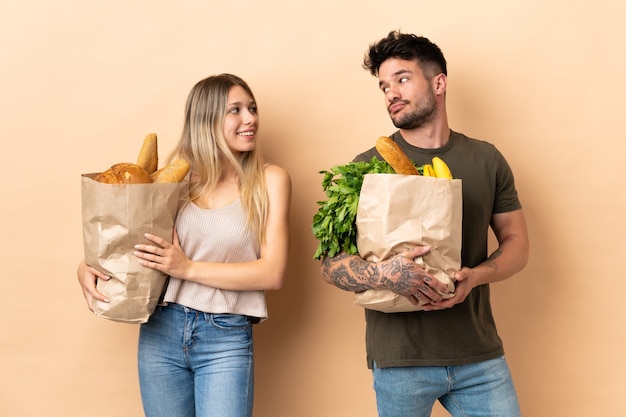 Coppie che tengono i sacchetti della spesa della drogheria sopra fondo isolato che osserva sopra la spalla con un sorriso