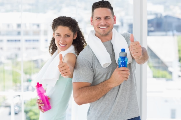 Coppie con le bottiglie di acqua che gesturing i pollici su alla palestra