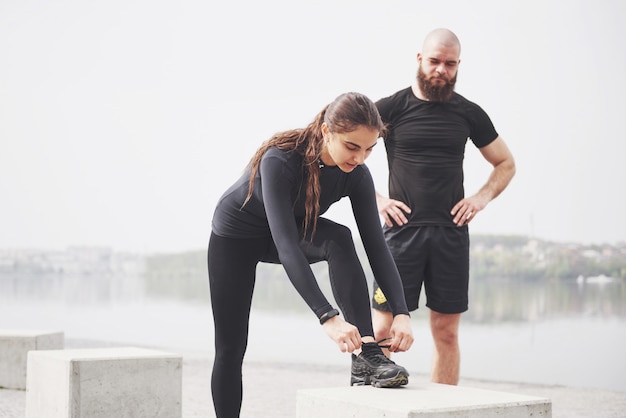 Coppie di forma fisica che allungano all'aperto nel parco vicino all'acqua. Giovane uomo e donna barbuti che si esercitano insieme nella mattina