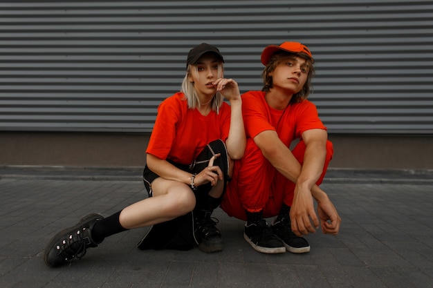 Coppie di modo con cappelli alla moda in vestiti alla moda arancioni con scarpe da ginnastica che si siedono vicino al muro di metallo grigio