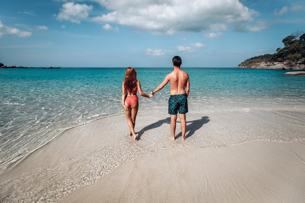 Coppie di vacanza che camminano insieme sulla spiaggia nell'amore che tengono le mani. Felice giovane coppia: uomo in pantaloncini e ragazza dai capelli rossi in un bikini rosso. Vista posteriore. La migliore vacanza. Phuket. Tailandia.