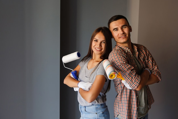 Coppie felici con il rullo di pittura che guarda alla macchina fotografica sulla parete grigia