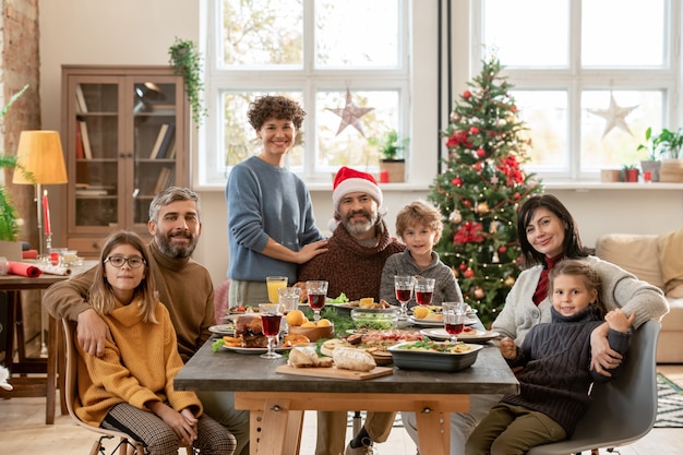 Coppie giovani e mature felici e tre bambini carini riuniti da un tavolo festivo servito per la cena di Natale in soggiorno contro un albero decorato