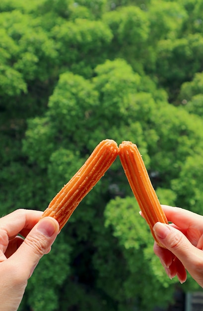 Coppie mani tintinnio bastone di churros fritti freschi contro fogliame verde sfocato