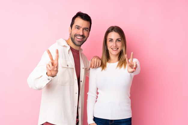 Coppie nel giorno di S. Valentino sopra la parete rosa isolata che sorride e che mostra il segno di vittoria