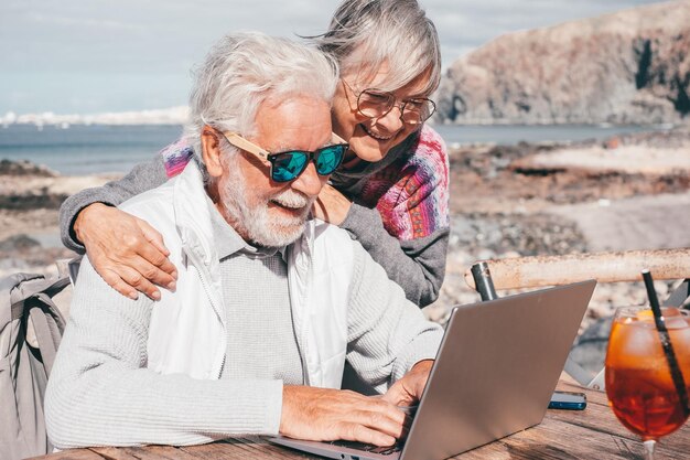 Coppie senior attraenti allegre che passano in rassegna insieme sul computer portatile che gode del giorno pieno di sole all'aperto alla spiaggia