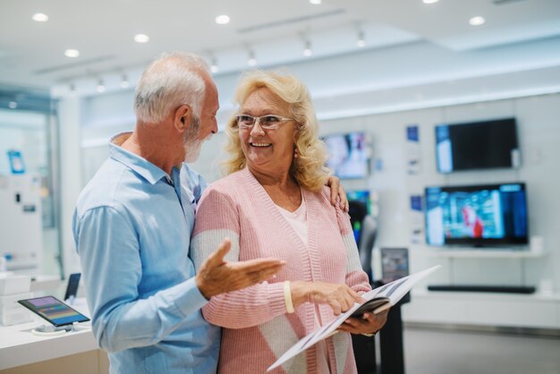 Coppie senior caucasiche felici che esaminano opuscolo e che scelgono che cosa comprare mentre stando nel deposito di tecnologia.