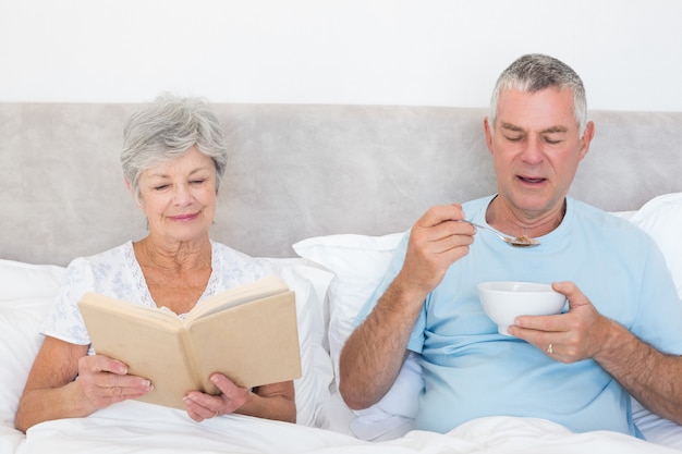 Coppie senior con il libro e ciotola di cereali a letto