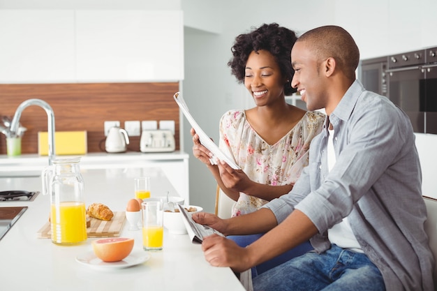 Coppie sorridenti che leggono insieme e che mangiano prima colazione nella cucina a casa