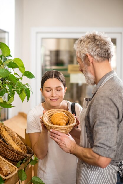 Cornetto profumato. Bella donna dai capelli scuri con gli occhi chiusi inalando aroma di croissant dal cesto nelle mani del venditore dai capelli grigi