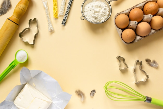 Cornice di ingredienti per la cottura di Pasqua e utensili da cucina. Farina, uova, burro e confettini dolci su fondo beige.