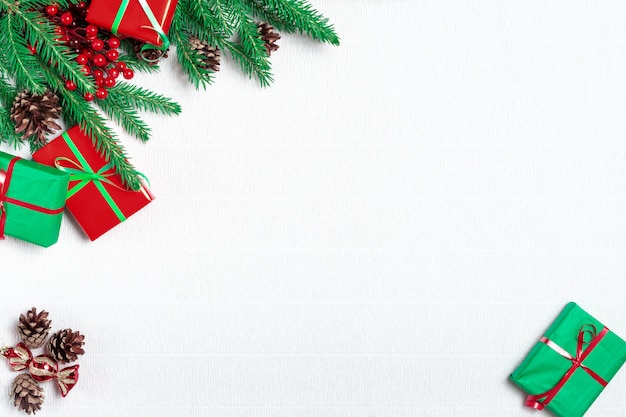 Cornice di Natale fatta di rami di abete, decorazioni festive, scatole regalo, bacche di agrifoglio rosso e pigne sul tavolo bianco.