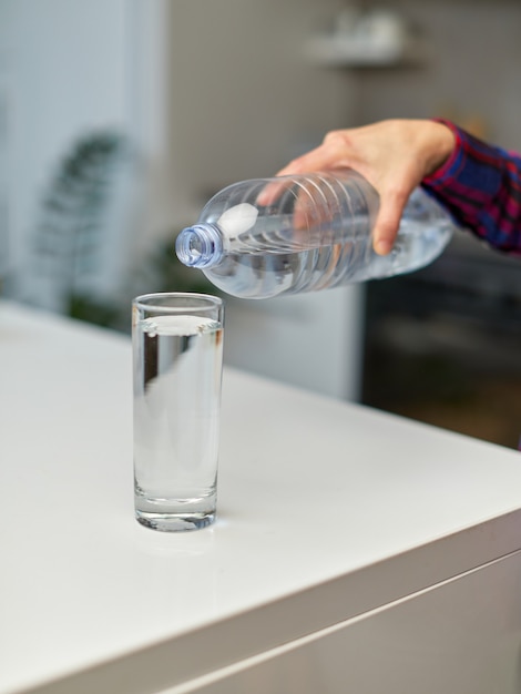 Cornice immagine di una mano femminile che tiene in mano una bottiglia di acqua potabile e versa acqua nel bicchiere sul tavolo su sfondo sfocato della cucina.